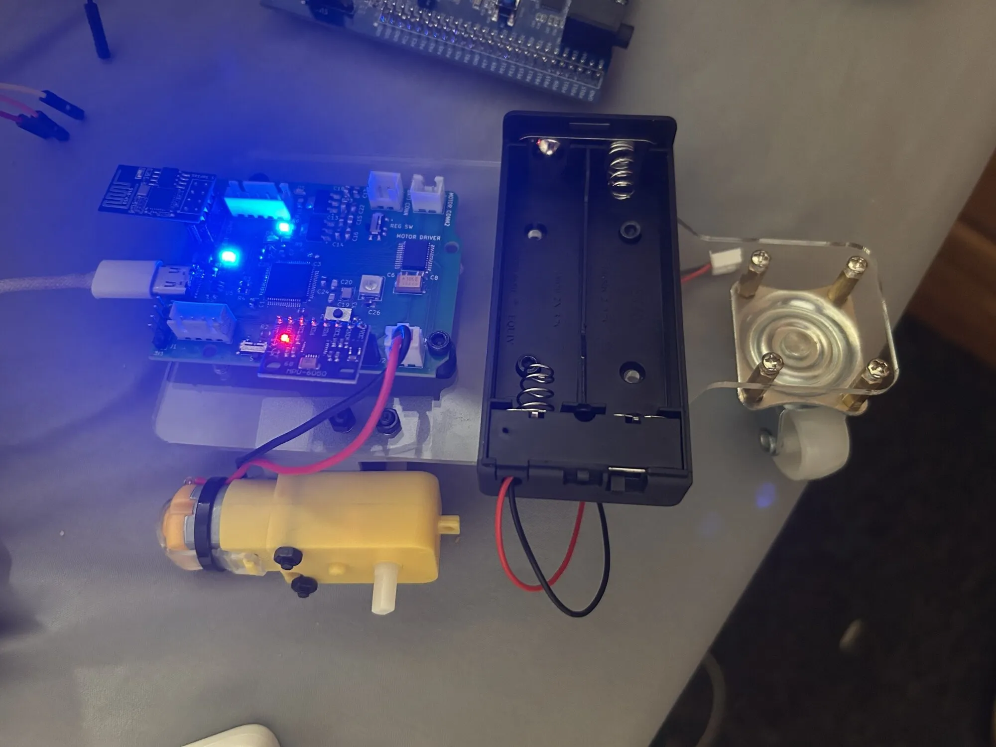A green robot controller board on a clear acrylic plate with a blue LED, an ESP8266 module and an IMU module lit, beside a battery holder, a yellow gear motor and a caster